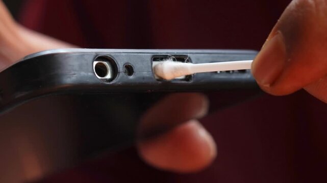 Close-up of an Asian man's hand cleaning a smartphone charging port with a cotton swab. Removing dust, lint, and debris from the charging connector for better device maintenance and smartphone care.