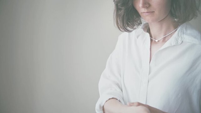 Young woman adjusting her clothing in a white button-up shirt and pearl necklace against a soft indoor background, showcasing an elegant lifestyle in close-up.