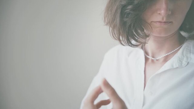 Young woman adjusting her clothing in a minimal indoor setting, wearing a white button-up shirt with shoulder-length wavy hair, lifestyle and beauty concept.