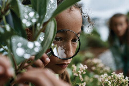 A focused young girl examines plants with a magnifying glass, merging curiosity with nature, while exploring the intricacies of the environment around her, fostering a love for the outdoors.