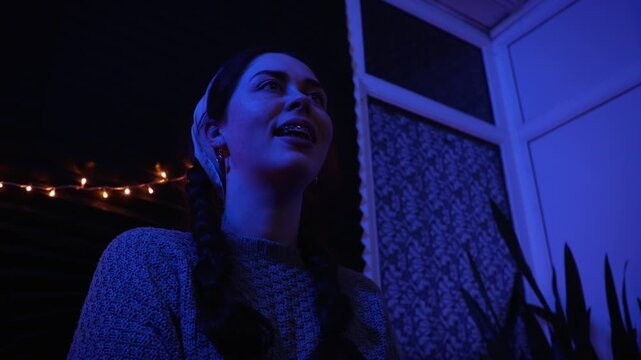 Young Caucasian woman sitting modestly at a party, gently moving to the rhythm of music and shyly greeting guests. Colorful party lights create a festive atmosphere