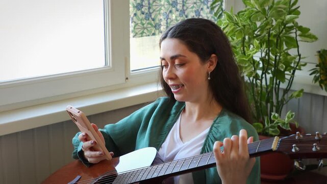 Portrait of a happy beautiful young Caucasian woman learning to play the guitar with online lessons on a smartphone. Modern remote education, digital learning, and self development concept.