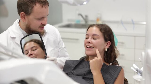 Interested young woman sits in a dentist chair and carefully looks at her teeth in the mirror. Male dentist with an assistant shows the client the work done. High quality 4k footage