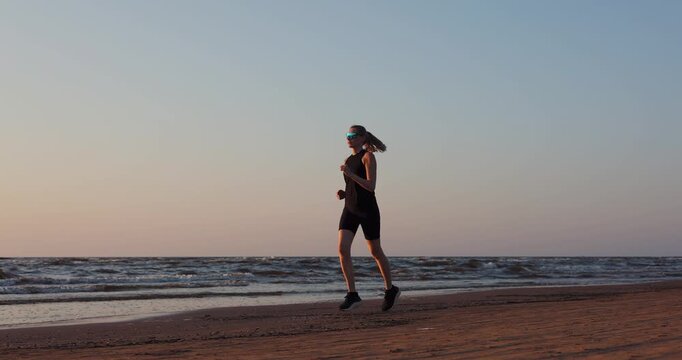 Sporty woman running on beach at sunset in black sportswear. Healthy lifestyle and fitness. Cardio workout
