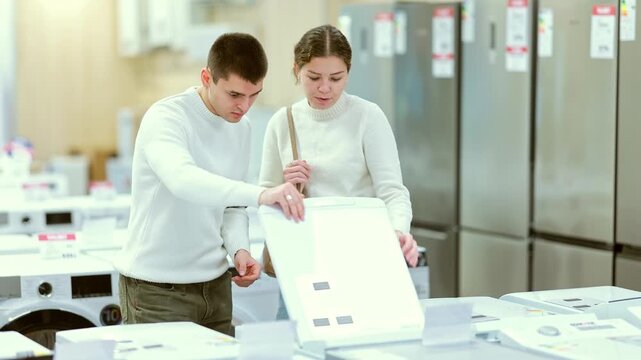 Couple customer inspect product range and selects dishwashing machine or washing machine. Customer chooses household appliances in store exhibition hall