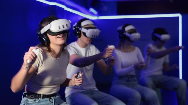 In darkened hall of gaming club, girl in VR helmet takes part in group game. Player overcomes obstacles and reaches goal in parallel reality. Imaginary fantastic world 