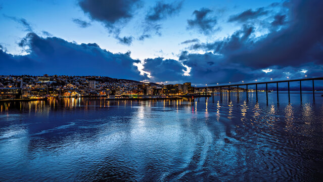 View from the water of the bridge and the waterfront promenade of Tromso, featuring numerous hotels. Blue hour at midnight in April.