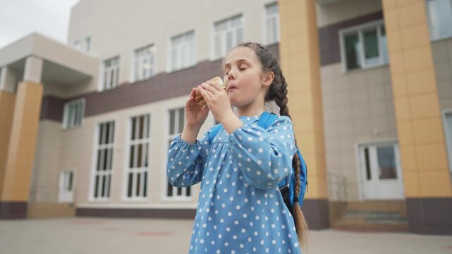 Schoolgirl eating sandwich outside school building. Child with backpack enjoys lunch near school. Girl with braid bites sandwich during break. Student eating food outdoors. Kid having snack at school.