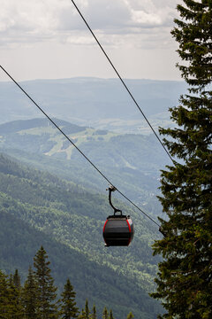 Cable car lift cabin moving on steel ropes above lush green mountain slopes covered with pine forest under overcast sky, mountain resort summer tourism and transportation concept