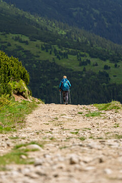 hiker with blue backpack and trekking poles climbing rocky mountain trail toward green alpine hillside, outdoor trekking adventure concept.