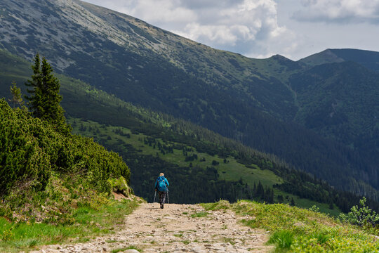 Lone hiker with blue backpack descending rocky mountain trail through green alpine valley, scenic outdoor trekking adventure concept.