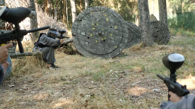  Tactical paintball team fighting on outdoor location. Men and women shooting with paintball markers, hiding behind barricade.