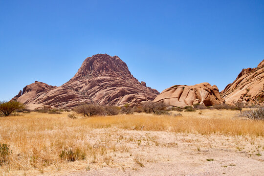 Eine Felslandschaft mit Granitberg, trockener Ebene und blauem Himmel zeigt Spitzkoppe in Namibia.
