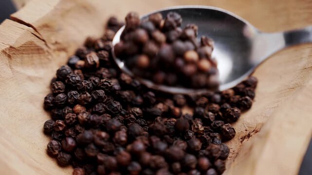 Stainless steel spoon scooping organic dried black peppercorns (Piper nigrum). Raw aromatic spices in rustic hand-carved wooden bowl