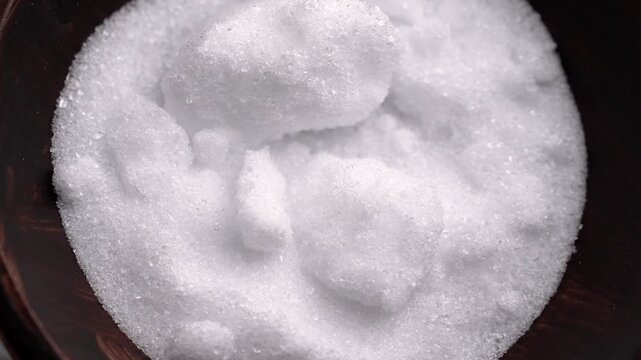 Metal spoon scooping Epsom salt crystals from a wooden bowl. Coarse white magnesium sulfate particles