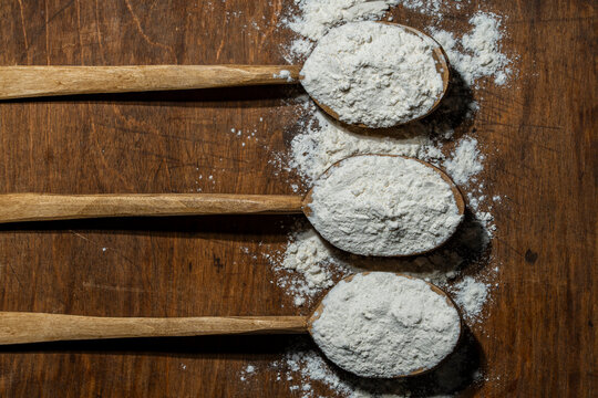 Wooden spoons with wheat flour on rustic dark background top view, closeup. Three wooden spoons filled with white baking flour on vintage board. Organic ingredient concept for bakery and cooking blog