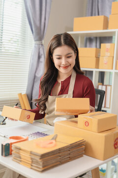 Smiling Asian woman entrepreneur holding pen and checking customer order on digital tablet while preparing parcel boxes at her home office desk.