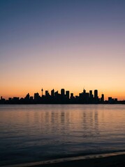 Fototapeta premium The image captures a stunning silhouette of a city skyline against a vibrant, colorful sky. A calm body of water reflects the buildings and the sunset, creating a mirrored effect.