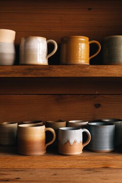 Ceramic mugs on wooden shelf. Handmade rustic cups for tea or coffee in pottery workshop. Traditional artisan craft for home interior and cozy kitchen design aesthetic collection.
