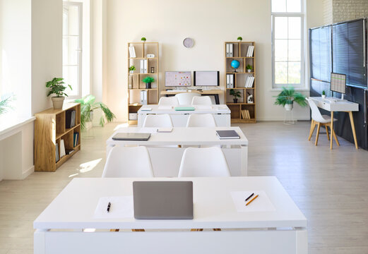 Modern classroom interior with empty desks for education. Front view with white tables, chairs and laptops in a bright school setting, ready for lessons and training. A neat study space idea.