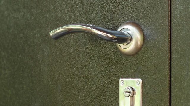 A close-up shot of a hand pressing a chrome metal handle of a heavy entrance door. A person smoothly opens a dark armored door to go inside the house or building.