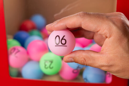 Gros plan sur une main tenant une boule de tirage au sort pour le loto, le num&eacute;ro 6. En arri&egrave;re-plan, une boite transparente avec des autres boules.