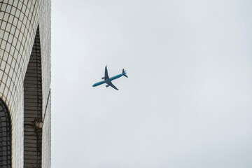 Minimalist urban composition featuring a cerulean blue airplane soaring past a white tiled skyscraper against a muted grey sky, evoking themes of travel and globalization. © Yu