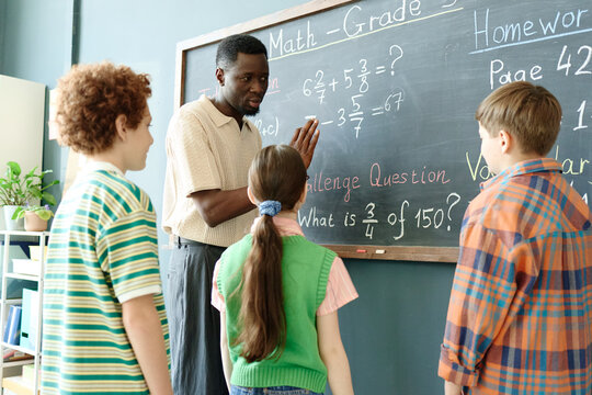 Black male teacher explaining math problem on chalkboard to elementary school children, guiding classroom learning and teamwork. Useful for education marketing, tutoring, school programs