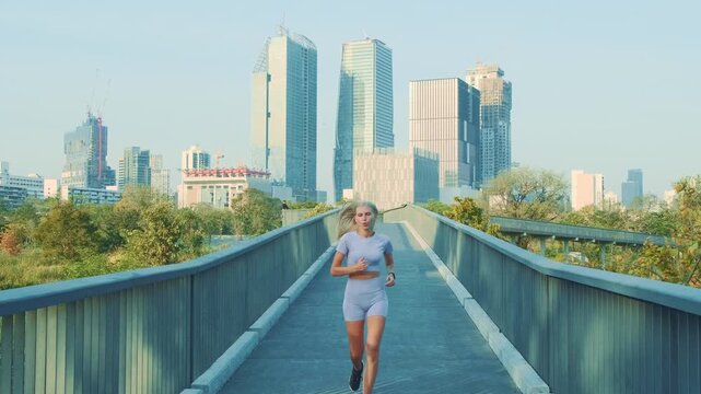 A young blonde woman runs at her own pace along a path in a green urban park in Bangkok, with skyscrapers and apartment buildings visible behind her.