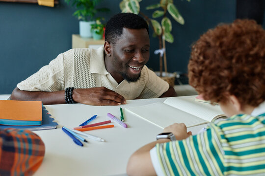 Medium shot of Black male teacher guiding schoolboy during writing practice at desk, supporting literacy and learning. Useful for education, tutoring, child development campaigns