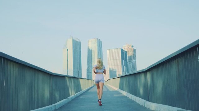 A blonde woman in athletic clothing runs alone at a comfortable pace on a pedestrian path at nighttime, with skyscrapers visible in the background.