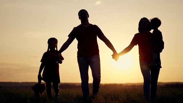 Family walks together at sunset holding hands across grass field. Parents with two children enjoy evening walk. Girl carries teddy bear. Mother holds son. Family silhouettes against sun.