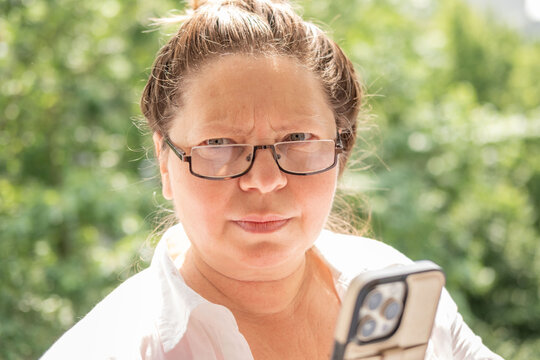 woman with glasses looking smartphone frustrated expression cyberbullying victim, online conflict, technology stress, furrowed brow, intense gaze, white shirt, green background, bad news, screen time