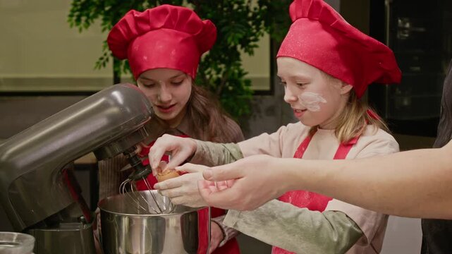 Caucasian teenager and child wearing chef hats cracking egg into stand mixer bowl during baking class. Footage ideal for culinary school promotions and family lifestyle advertisements