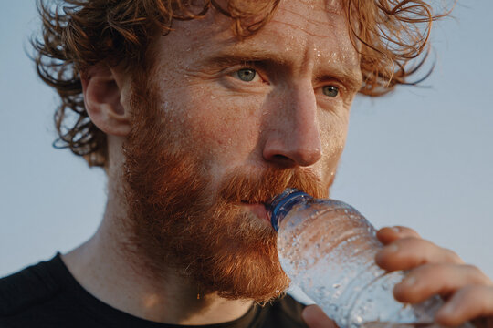 Sweaty redheaded man drinking water from plastic bottle after outdoor workout, focused recovery and hydration in warm natural light