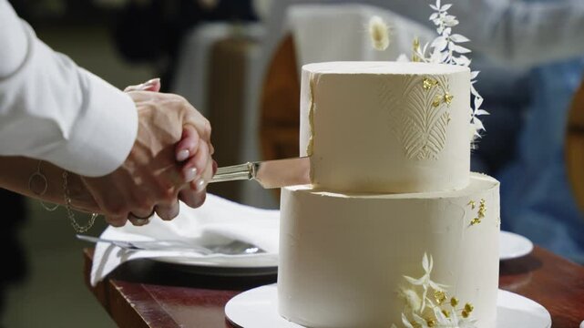 Hands hold a knife to cut a wedding cake adorned with flowers during a reception, with guests and tables in the background