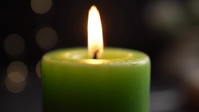 Burning green cylinder candle with a glowing flame in a dark room with soft bokeh lights