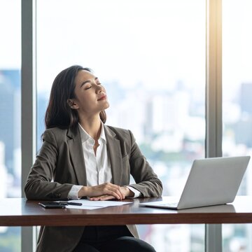 A peaceful Asian businesswoman takes a deep breath of relief, enjoying a calm, serene moment of mindful meditation and success at her bright city office desk with a laptop.