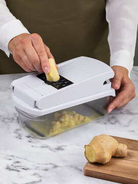 Woman grating fresh ginger on manual vegetable slicer with grater attachment on marble table closeup