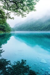 Obraz premium Serene blue lake reflecting misty mountains and lush green trees