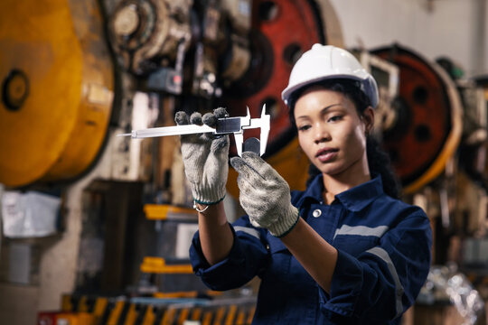 Confident female manufacturing engineer performing precision quality inspection using a digital caliper to measure a machined metal component in an industrial factory.
