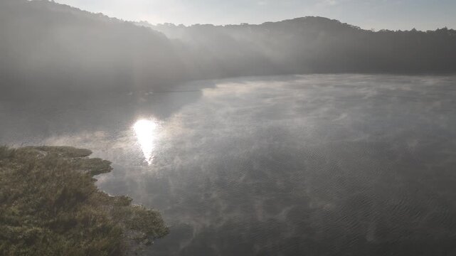 Beautiful cinematic footage of misty Tuyen Lam Lake covered in thick fog during morning sunlight in Dalat, Central Highlands, Vietnam. A serene nature landscape with sun rays reflecting on the water