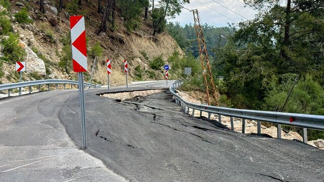 Mountain road damaged by landslide with cracked asphalt and bent guardrail, warning chevrons and forested hillside in view, conveying hazard and disruption