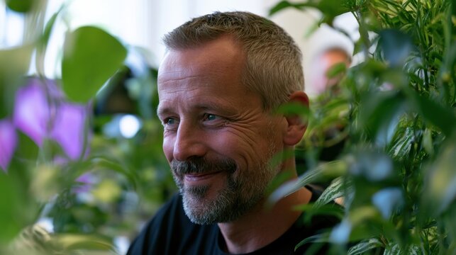Middle-aged man with short gray hair and beard smiling happily surrounded by green leafy plants indoors in soft natural light with blurred background