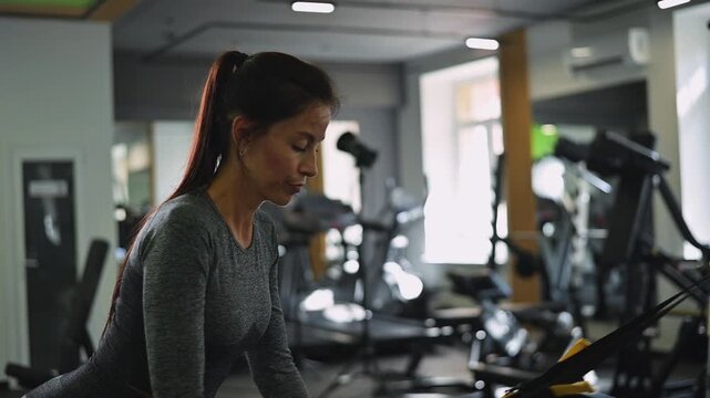 A woman in her late twenties performs strength training exercises in a gym. She uses resistance bands to engage various muscle groups while focusing on her technique and posture