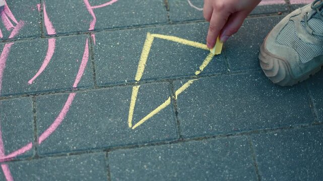 Woman drawing a yellow triangle on asphalt with a piece of chalk. Creative child play outdoors on a sidewalk. Educational game for kids and artistic hobby.