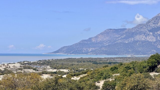 Coastal landscape with sandy dunes and green shrubs beside a calm blue sea, distant mountains under clear sky, peaceful natural scenery for travel and nature themes