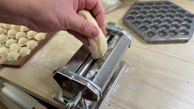 Hands feed dough into a metal pasta machine on a wooden counter, flour dust and small dumplings nearby, warm kitchen scene suggests homemade cooking