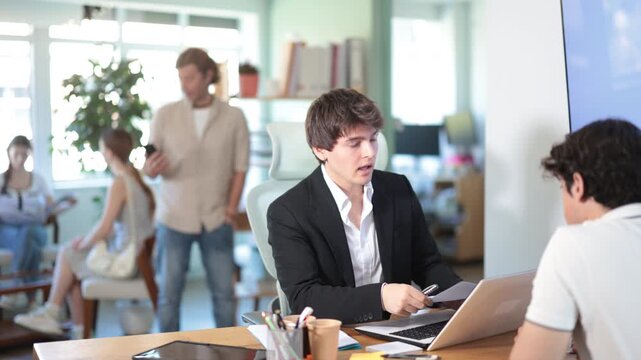 Adult male bank employee sits at desk with laptop and discussing with young male customer 