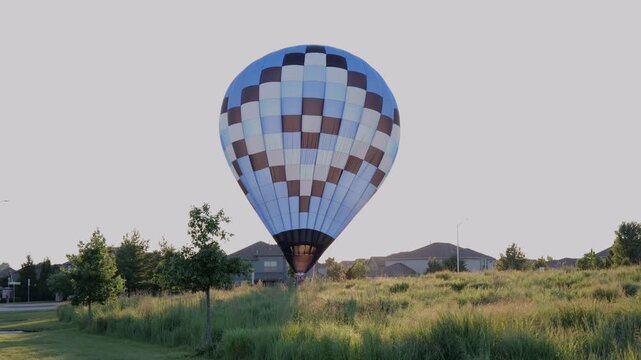 Large blue and brown checkered hot air balloon touching down and deflating in a grassy field near a suburban neighborhood at sunset